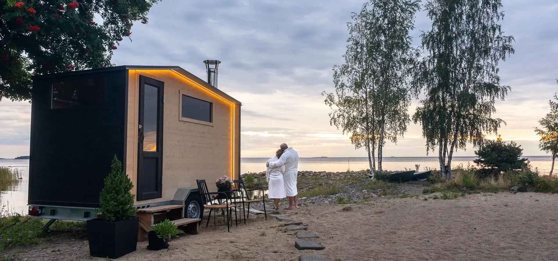 Pariskunta nauttii juhannussaunasta hiekkarannalle sijoitetun kotimaisen Lapelland Saunavaunu S -mallin terassilla.
